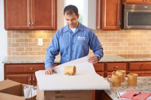 An Olympia mover in a blue shirt carefully wraps mugs in paper, on top of a moving box, in a kitchen, representing quality relocation services by an Olympia Moving Company since 1995.