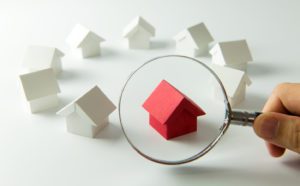 A hand with a magnifying glass examining a red model of a house, amongst a circle of eight models of white houses, representing how to find the perfect home to buy.