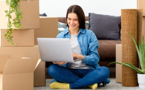 A woman sitting on the floor surrounded by moving boxes, using a laptop, in Washington, demonstrating how to go about choosing the right moving company.