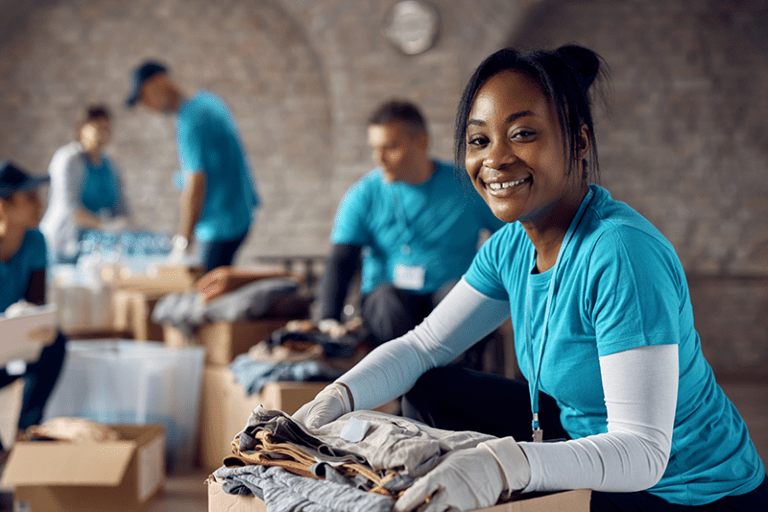 A woman in a blue shirt and gloves smiles while holding a moving box filled with folded clothes, demonstrating that Olympic Moving & Storage is proud to be part of the community.