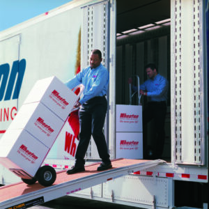 Two interstate and long distance movers load boxes into a moving truck, using hand trucks and the truck's ramp, demonstrating transportation services.